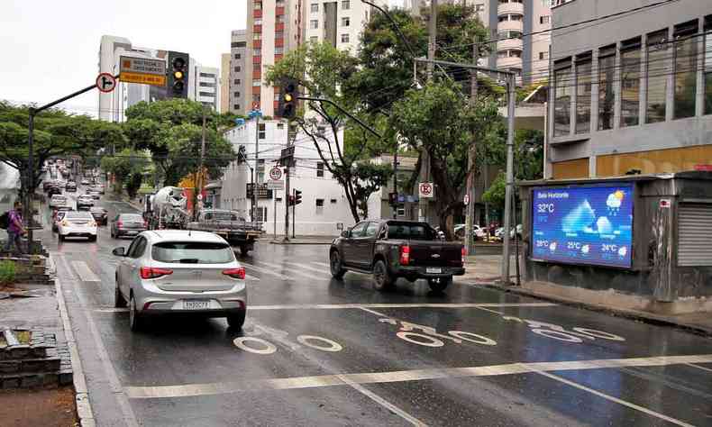 Rua da região central de Belo Horizonte com asfalto molhado. Carros estão parados no sinal. Há uma banca de revista exibindo a previsão do tempo.