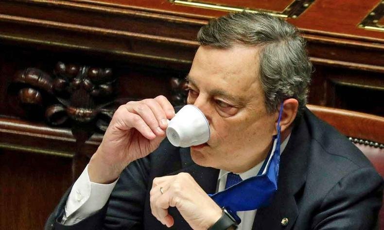 Mario Draghi, com m�scara abaixada, sorve caf� de uma x�cara branca, sentado numa cadeira do parlamento italiano