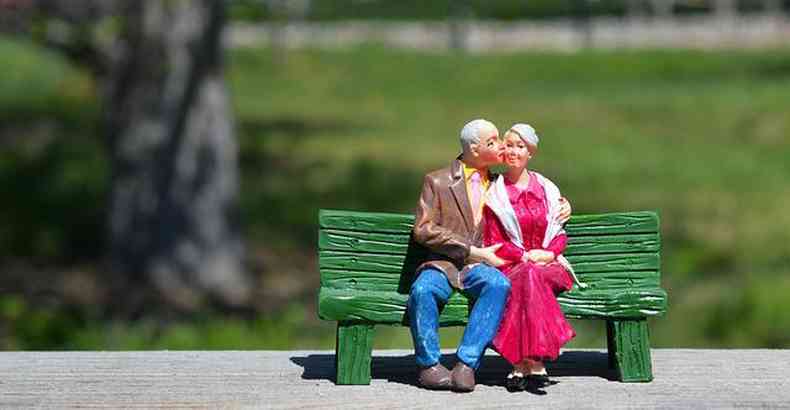 bonecos de um casal de idosos em banco de praça, o homem beijando o rosto da mulher