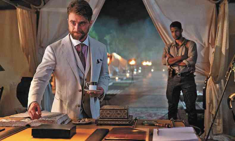 O ator Daniel Radcliffe, de terno branco, segura xcara de caf e tem a outra mo sobre um livro, observado por um homem ao fundo, na cena do filme Cidade perdida 