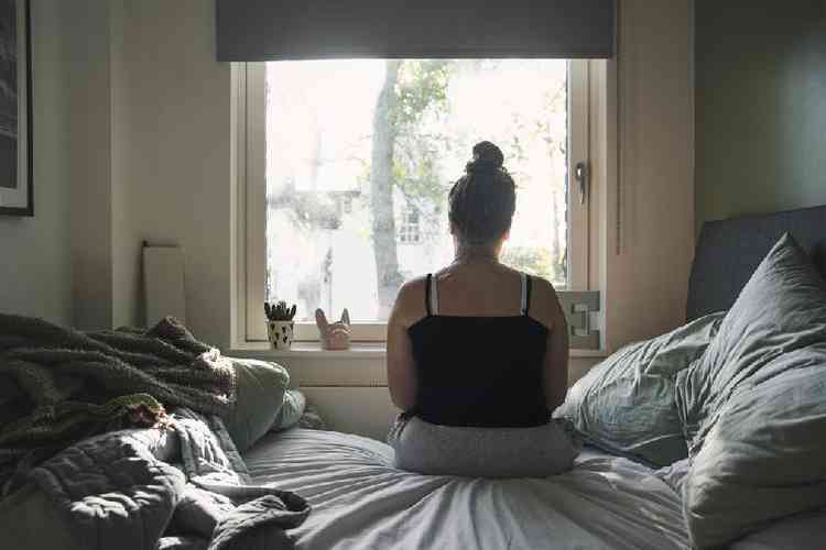 jovem mulher de costas sentada na cama e olhando pela janela