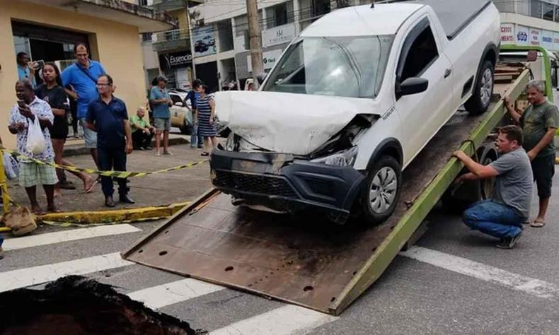 Caminhonete saveiro branco com a frente destruda sendo guinchada, pessoas ao redor vendo a movimentao