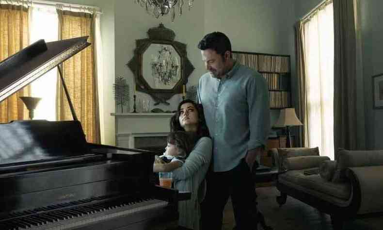 sentada ao piano segurando um beb, Ana de Armas olha para Ben Affleck, de p, atrs dela 