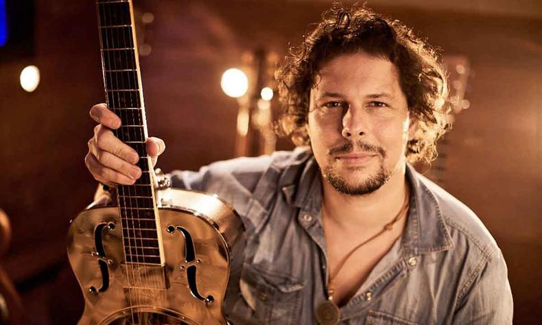 Ao lado do violo, msico Guilherme Schwab sorri, tendo ao fundo a luz de um refletor 