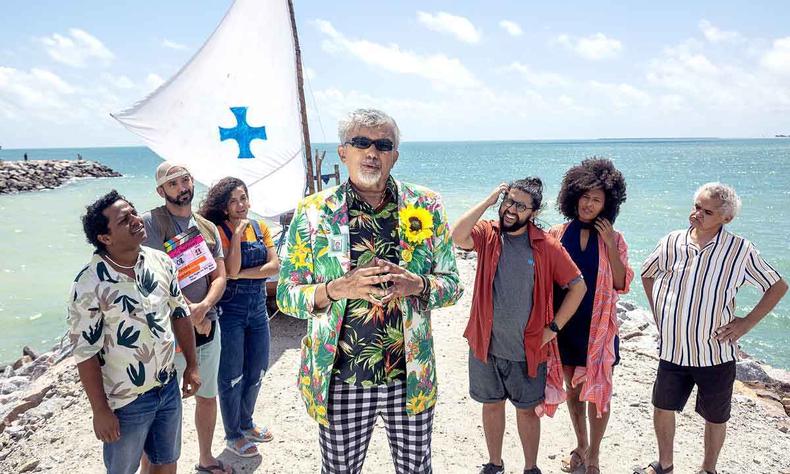 Humorista Falco est no centro de grupo de sete pessoas. No fundo, vem-se o mar e o cu azul