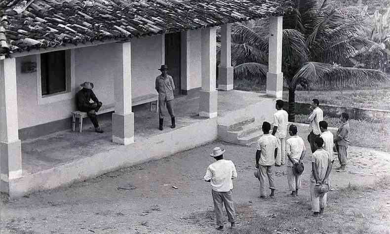 Homens esto na varanda de uma fazenda e,  frente deles, sob o sol est um grupo de camponeses