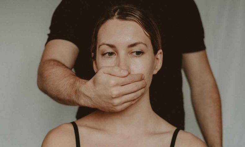 O silenciamento tamb Mulher branca com camiseta preta com um homem tampando sua boca por tr