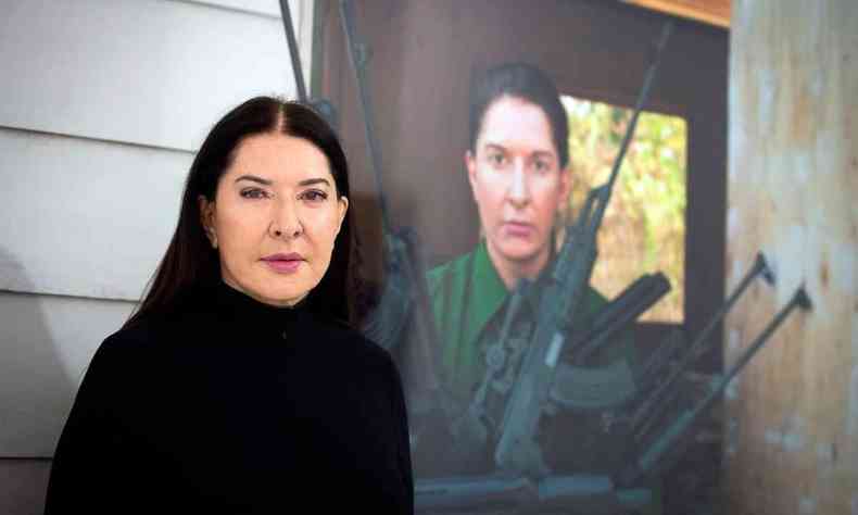 Artista srvia Marina Abramovic olha para a cmera. Ao fundo, v-se quadro com ela carregando armas 