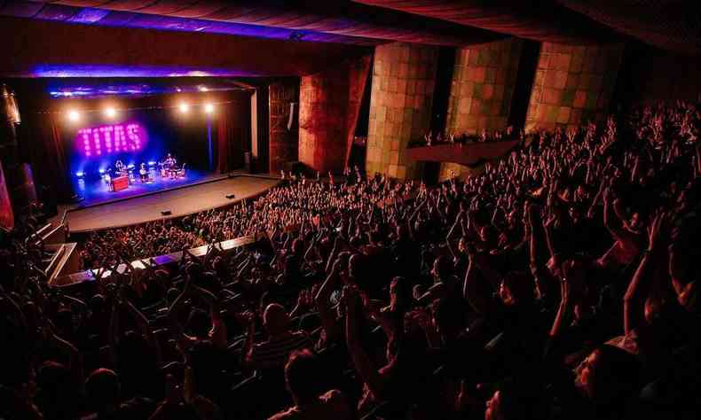 Foto mostra o Palcio das Artes lotado durante o show dos Tits. No palco, imenso telo exibe o nome da banda