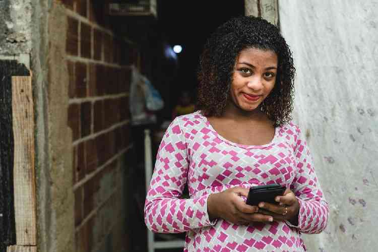 Mulher jovem de baixa renda segurando um celular e sorrindo timidamente para a cmera