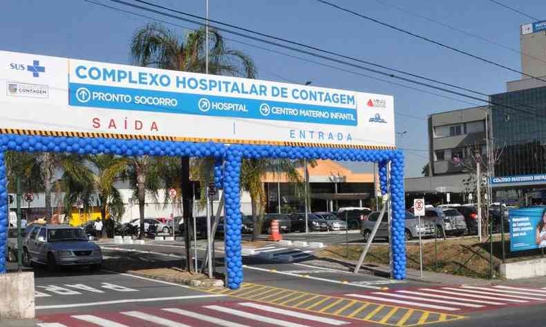 Entrada/portaria do Complexo Hospitalar de Contagem com grades, cancelas e letreiros em fundo azul
