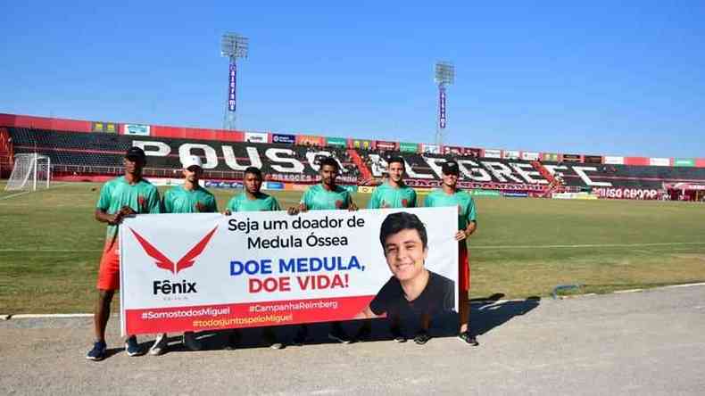Jogadores do Pouso Alegre entram em campo no Manduzo com faixa pedindo doao; na faixa aparece a foto de Miguel