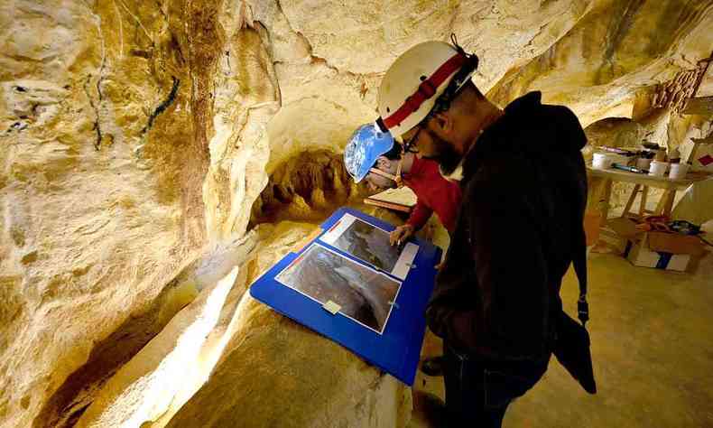 Homens de capacete veem fotos de pinturas rupestres, pregadas em cartolinas azuis. Eles esto dentro de caverna onde as imagens foram pintadas nas paredes