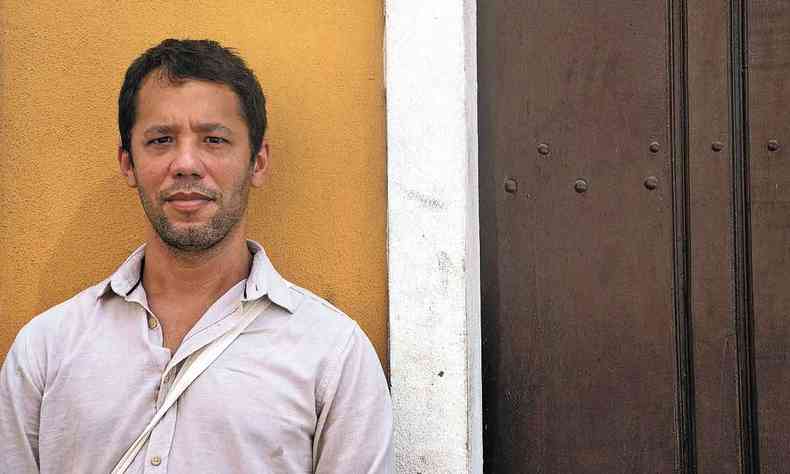 De camisa branca, Itamar Vieira Junior posa para foto, de p, encostado em parede amarela