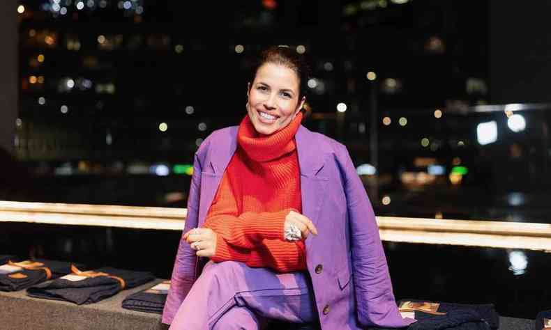 Usando blusa vermelha e terninho roxo, Flavia Soares sorri, sentada, tendo ao fundo a cidade à noite