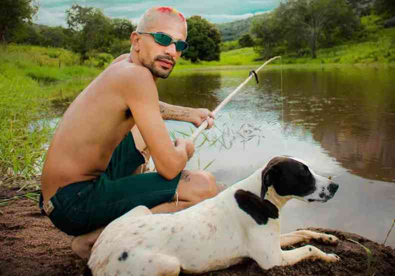 De bermuda, sem camisa e com culos escuros, rapper Oreia segura vara de pescar na beirada de um rio, com um cachorro branco com manchas pretas sentado ao seu lado