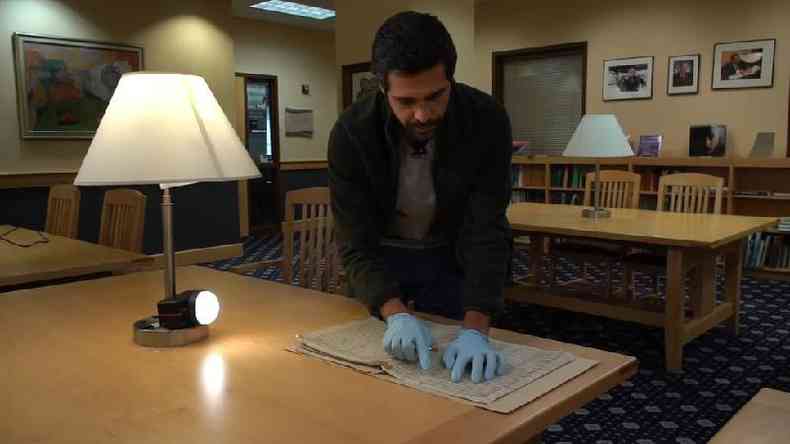 Franco Galvo em p, debruado diante de mesa, manipulando partituras com luvas em ambiente de biblioteca