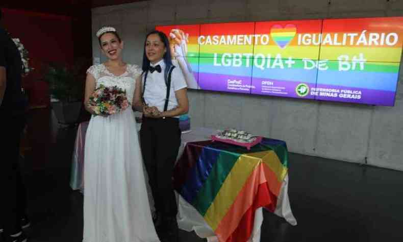 Duas mulheres posam para a foto olhando para a cmera. A da esquerda veste um vestido de noiva branco e segura um buqu de flores e a da direita usa camisa e cala social com um suspensrio. Ao fundo, um painel com as cores do arco-ris onde se l: 'Casamento igualitrio LGBTQIA+ de BH'