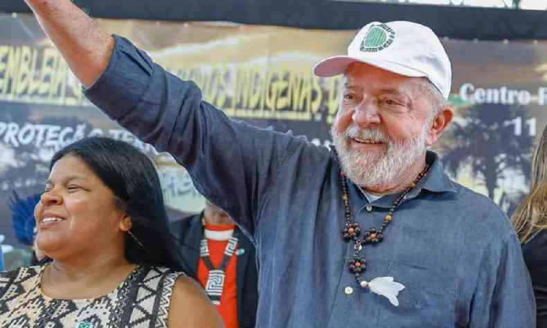 Lula com camisa azul escura e bon branco acenando com o brao direito ao lado de Snia Guajajara