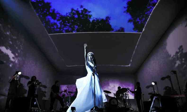 Marisa Monte, de vestido branco no palco, levanta a mo diante do microfone. No teto retrtil, veem-se o cu da noite e sombras de rvores. Ao lado dela, sombras dos instrumentistas