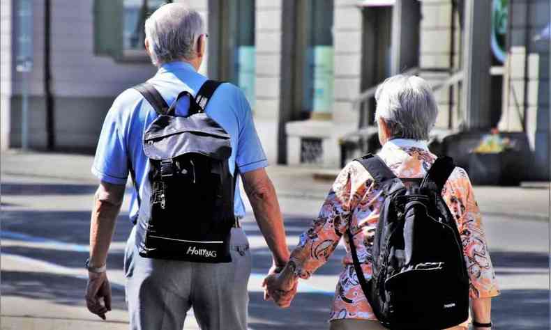 Casal idoso, com mochilas nas costas, caminha de mos dadas e de costas para a cmera 