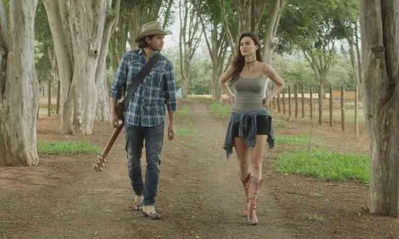 Atores Gabriel Sater, segurando o violo, e Taila Ayala caminham por estrada no filme Corao de cowboy