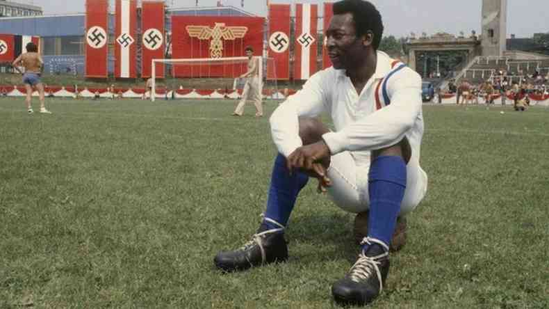 Pel com roupa branca e meio azul sentado no campo de futebol, atrs, bandeiras nazistas
