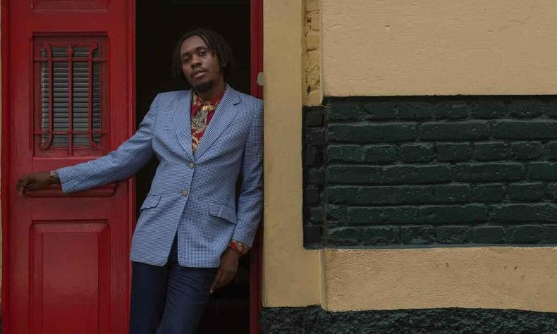 Rapper Rincon Sapiencia posa em frente a uma porta vermelha, escorado em parede amarela com detalhes em preto