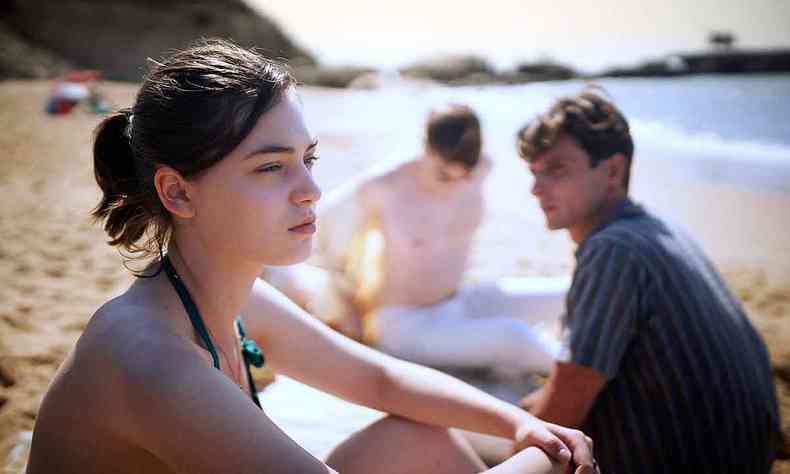 Atriz Anamaria Vartolomei de biquni, sentada na areia da praia, com semblante srio, ao lado de dois rapazes com semblante srio