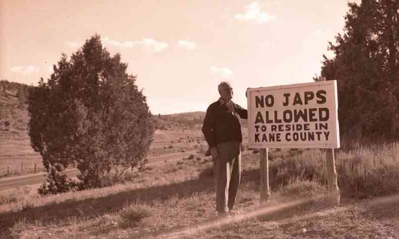 Homem branco parado em frente a um campo ao lado de uma placa com os dizeres 'No japs allowed to reside in Kane County', traduzido para 'Japs proibidos de residir no Condado de Kane'