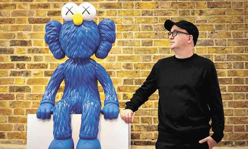 Artista plástico Kaws, de roupa preta, posa ao lado de escultura azul exposta por ele em galeria de Londres