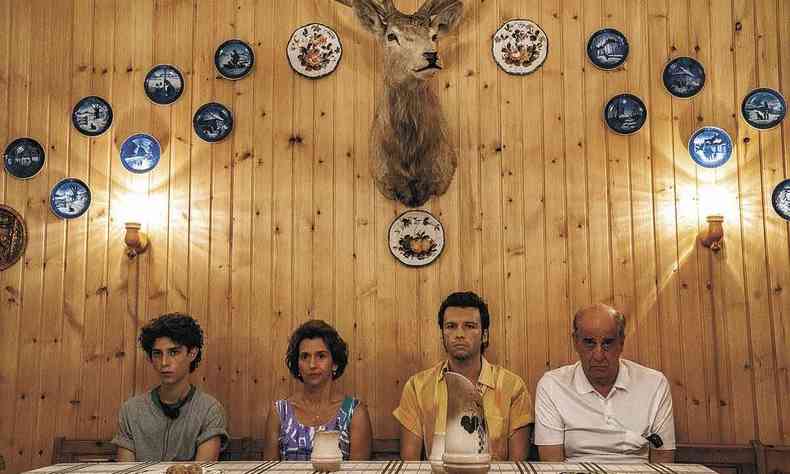 Sentados  mesa com semblantes srios, quatro integrantes da famlia Schisa tm ao fundo parede com pratos pendurados e enorme cabea de veado 