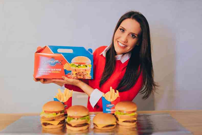 Mulher com blusa vermelha e cabelo preto sorri para foto, enquanto segura caixa de embalagem do hambrguer da marca. Quatro sanduches esto na sua frente, sobre a mesa.