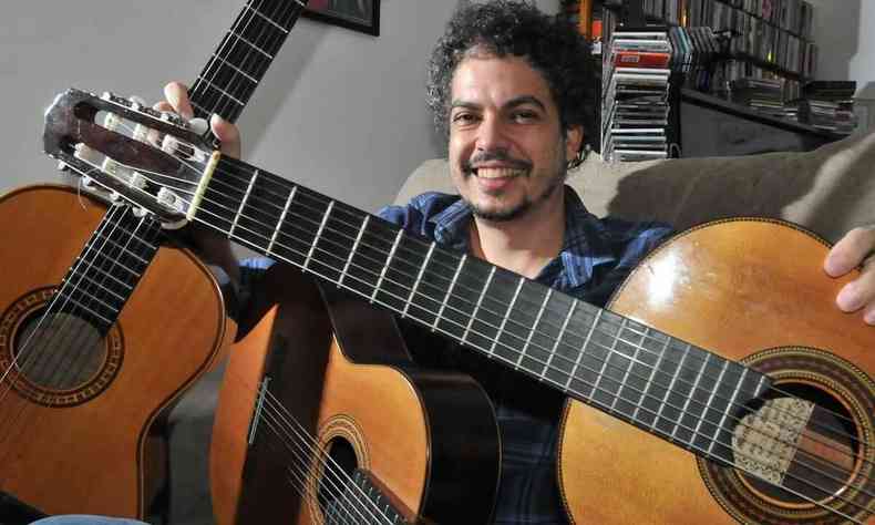 Thiago Delegado sentado na sala de sua casa abraça três violões e sorri