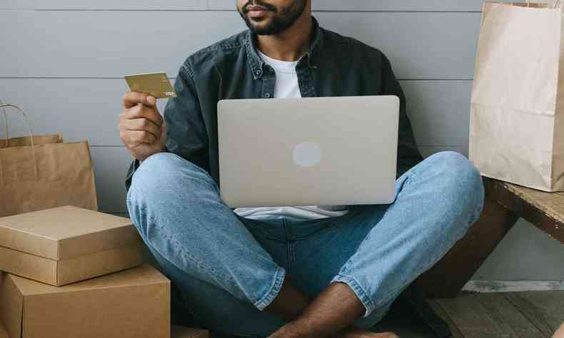 Homem sentado no chão segura cartão de crédito e notebook