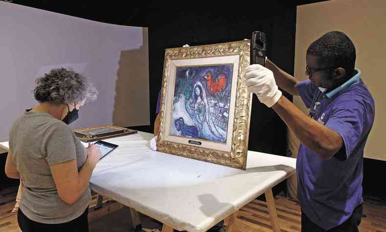 Usando luvas, técnico segura quadro de Marc Chagall, enquanto especialista toma notas sobre a pintura em um tablet 