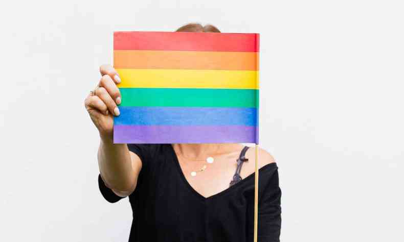 Mulher usando uma blusa preta segurando a bandeira LGBT a frente do rosto 