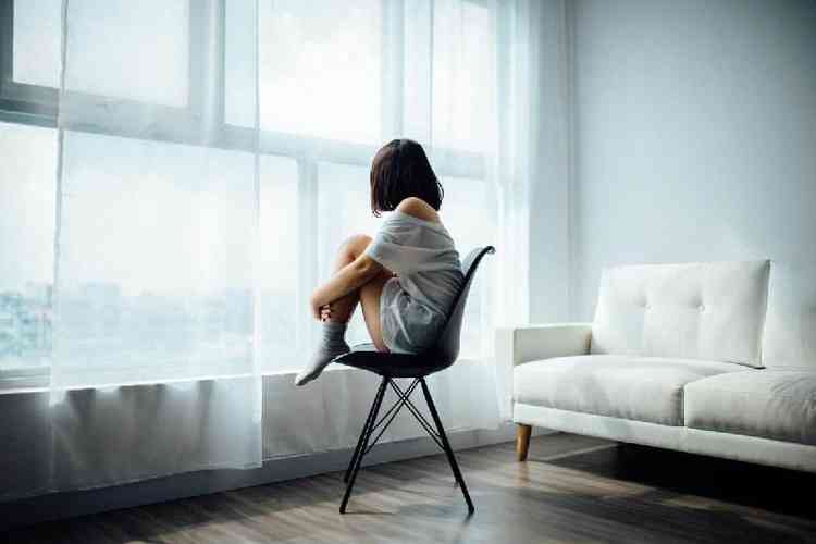 Mulher sentada, encolhida, diante da janela de um quarto