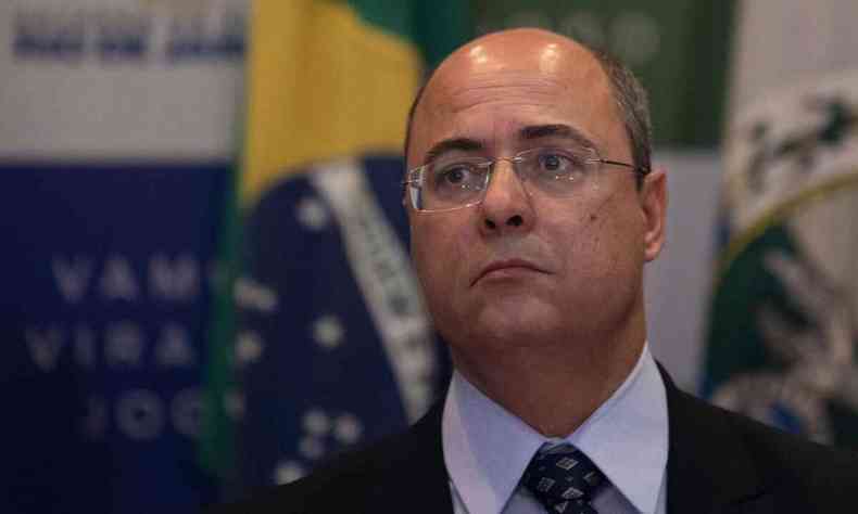 Wilson Witzel, ex-governador do Rio, é um homem calvo, usa óculos; ao fundo, vê-se a bandeira do brasil