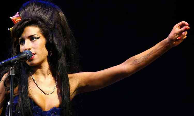 No palco, Amy Winehouse canta ao microfone e estende o brao esquerdo para o lado
