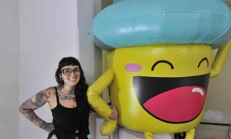 A artista Raquel Bolinho ao lado de seu personagem, cupcake sorridente de corpo amarelo e chapu verde 