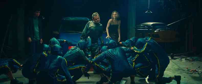 O ator Matheus Nachtergaele tem atrás de si uma jovem loura e está à frente de homens agachados, vestidos de preto, numa oficina de carros 