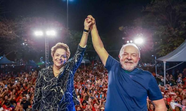 Dilma e Lula de mos dadas. Dilma a esquerda, uma mulher branca de cabelos curtos que usa uma camisa preta com listras brancas. Ao lado direito, Lula, homem branco de cabelos brancos com uma camisa azul