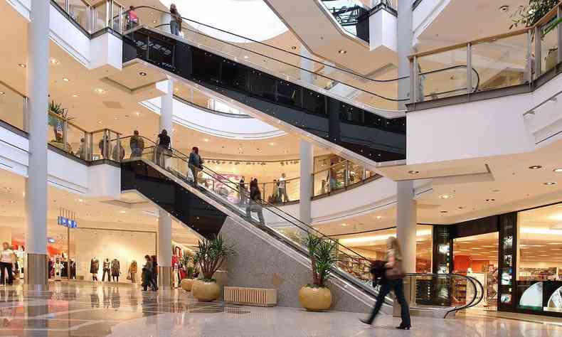 Imagem em tons de branco e iluminao clara, com duas escadas rolantes, lojas e pessoas, como em um ambiente de shopping.