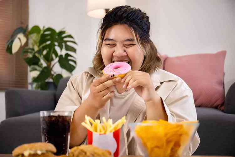 Menina asitica morena com sobrepeso em frente a uma mesa cheia de alimentos de fast food comendo uma rosquinha