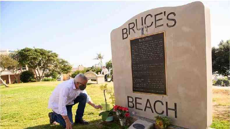 Homem coloca flores perto de placa em homenagem a Willa e Charles Bruce, em dia ensolarado