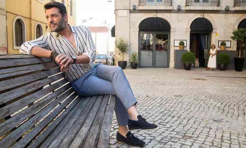 Rodrigo Lombardi, usando blusa listada branca e azul, est sentado numa praa em Portugal e olha para o lado