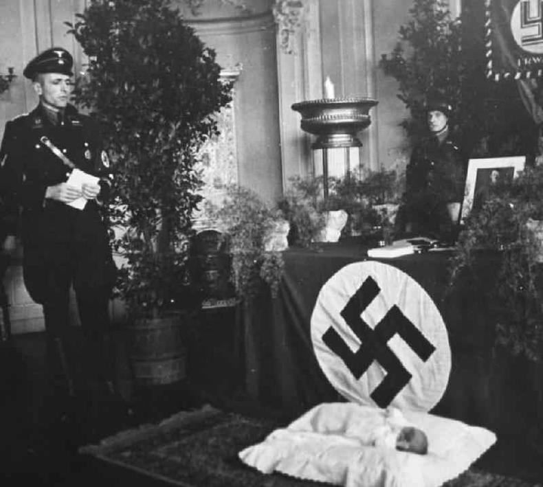 Oficial nazista olha para um criana deitada em bero em frente a uma bandeira com sustica