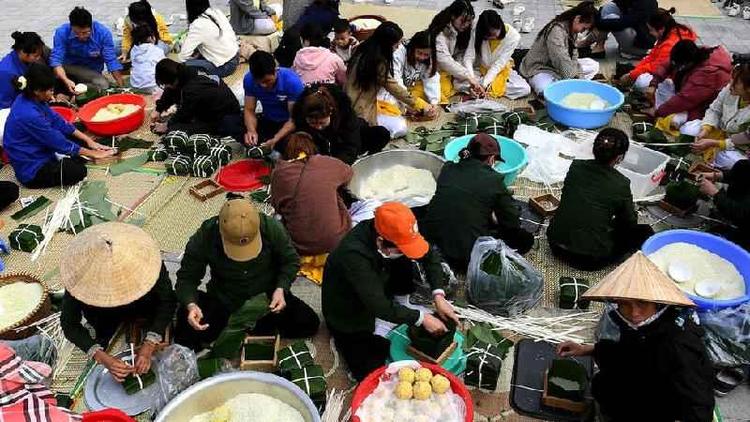 Um grande grupo de pessoas sentadas em esteiras fazendo bolos de arroz