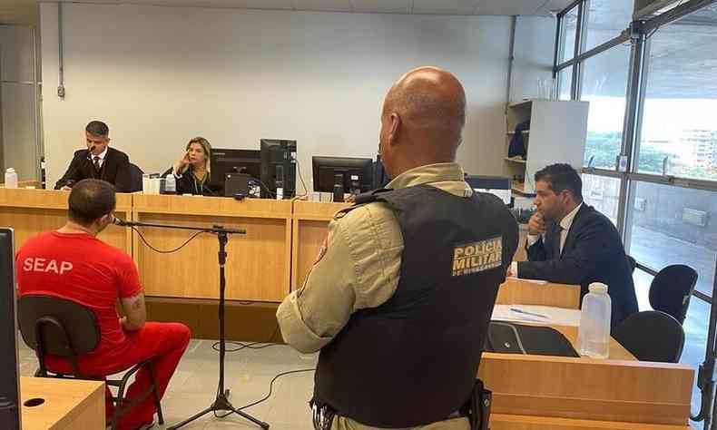 Julgamento do ru. Um policial est fardado de costas para a cmera, olhando para um rapaz sentado, vestindo roupas vermelhas. Juzes ao fundo.